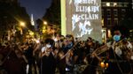 A photograph shows the violin vigil held on June 29, 2020, in New York City. Thousands line the street playing their instruments to honor Elijah, who was also, a violinist. In the center of the crowd is a sign with illuminated white doves, and lettering that reads: "In Memory of Elijah McClain". (Photo by Byron Smith/Getty Images)