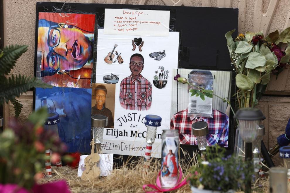 Photograph of a memorial for Elijah McClain, across the street from where the incident occurred. The image shows a multitude of objects placed to remember Elijah. Photographs and drawings of his image are surrounded by flowers, candles, patio lights and other objects.  on the bottom of the biggest poster are the words "Justice for Elijah McClain" . Below is another sign that reads #I'mDifferent