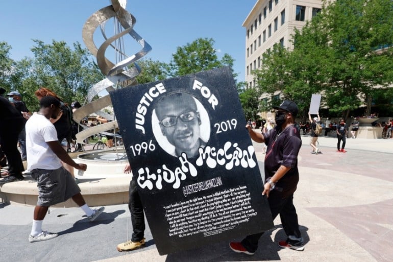 Photo taken during the 2020 protests, demanding justice in the death of Elijah McClain. Activists carrying signs march in the streets. In the foreground two protesters are carrying a huge sign with a picture of Elisha and text that reads: "JUSTICE FOR ELIJAH MCCLAIN – 1996-2019" at the bottom, above his final words appears text reading: #JusticeForElijahMcClain