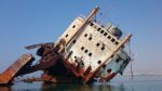 Photograph shows the remains of a large ocean vessel leaning as it rests in shallow water. The hull of the ship is rusted, tattered and demolished. All that remains are rusted beams and scraps metal dangle from the upper deck's carcass. The image symbolizes the poor and disabled "huddled masses", as well as the American political system. Time wasted on bickering and bureaucracy, has taken a toll in many ways.