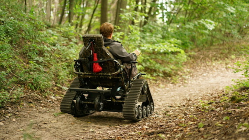 Image shows the rugged "TrackMaster" ATV wheelchair. Triangular-shaped tank style treads propel the vehicle throughout Rocky Forest Trail in Michigan. The drivers right hand is holding the joystick controller, which provides for easy maneuverability. Image courtesy of: "Bridge Michigan"