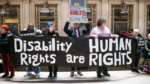 Photograph protesters holding a large banner that reads: "Disability Rights Are Human Rights". One protester holds a sign painted as a prison door showing only the top of the person's head to the barred window. The words "GUILTY!" In red lettering also appear on the sign.