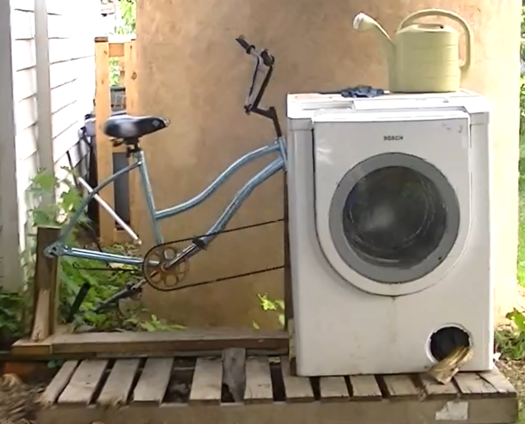 Image from footage taken by OutOfExile_IDR showing a pedal powered laundry alternative. A frontloading washing machine, is attached to the petal sprocket of a light blue stationary bicycle.