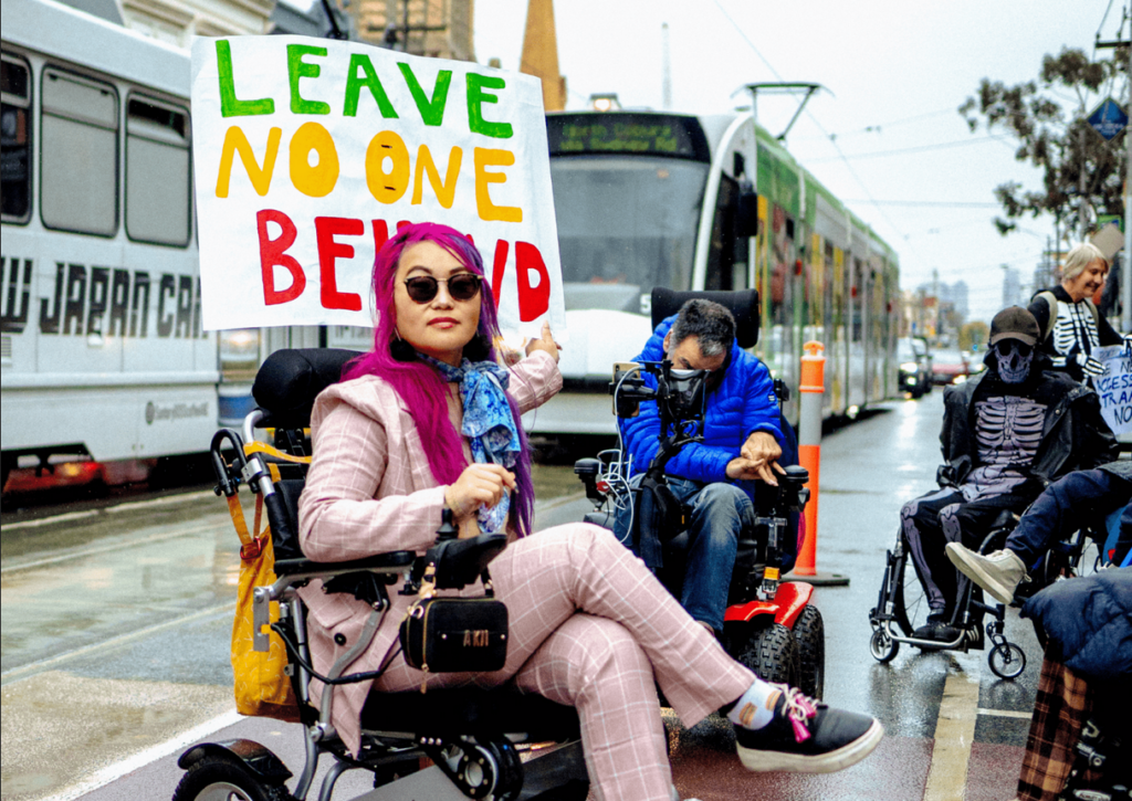 Image shows a number of disabled wheelchair user's on the street. A woman dressed in a light pink checker patterned suit, with pink and purple hair, points to assign reading: "LEAVE NO ONE BEHIND".
Image credit: Credit: © Ed Gorwell https://www.instagram.com/edgorwell/