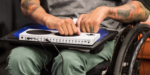 Photograph shows a wheelchair user who seems to have limb difference limitations. The person is wearing olive green pants and a gray shirt, with several arm tattoos. They appear to be manipulating the pad controls on the "Xbox Adaptive Controller" with the knuckles on their closed hand.