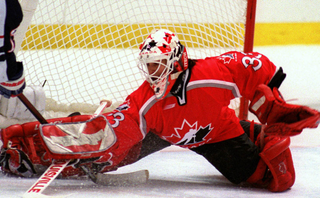 Photograph of Menell Rhéaume in 1998 in net for team Canada. She is stretching back across the net with her pad and blocker, poke checking the puck from the stick of the opposing US player. Image courtesy of Olympic CA