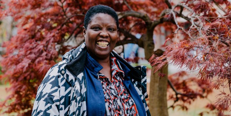 Image of African-American Woman Hero for Invisible Disability Rights, Lois Curtis wearing a multicolored shirt, a blue and white jacket and her customary big smile. Behind her is a tree with vibrant red leaves.